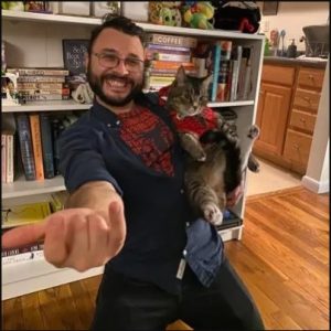 Dr Michael Tremmel is holding a cat while posing like Spiderman. He is smiling. He has a beard and glasses and a spiderman t-shirt is poking through his overshirt.