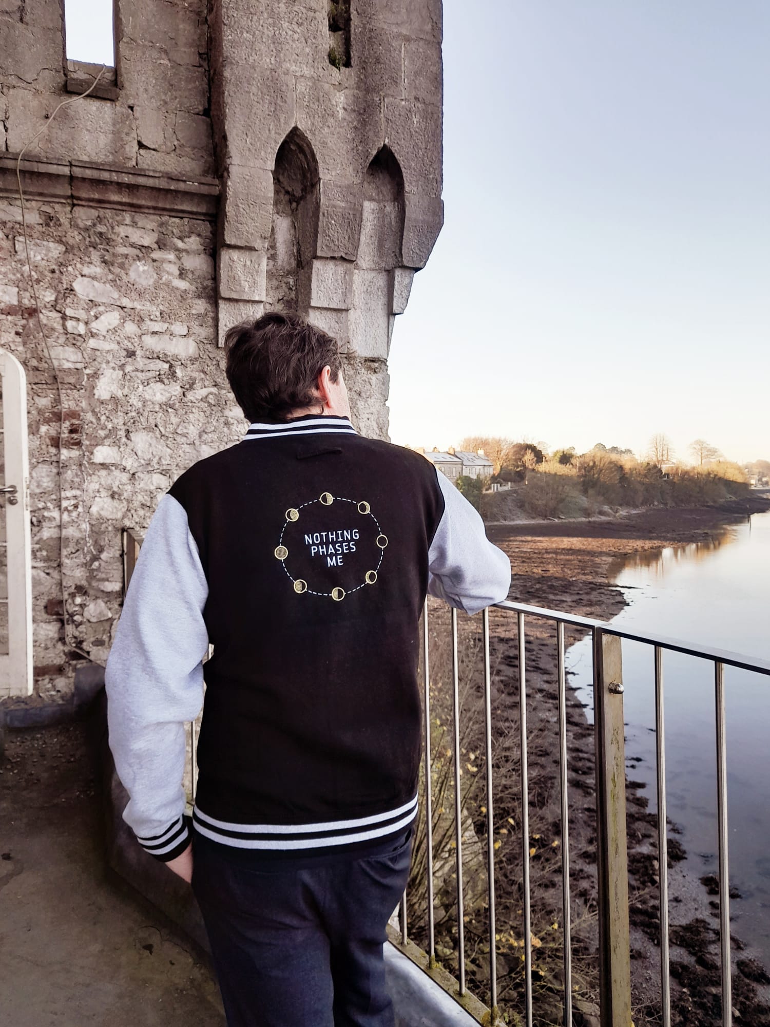 A man wearing a varsity style jacket with his back to camera. The phrase "Nothing phases me" is printed on the back of it along with a graphic displaying the phases of the moon. He is standing in front of a tower.