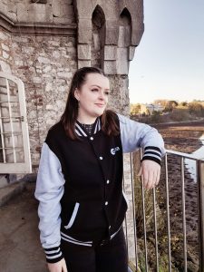 A woman wearing a varisty style jacket in Blacm and light grey. She is leaning on a railing and has a tower behind her.