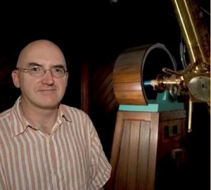 A photo of Professor Paul Callanan. He is standing in front of some large device constructed of wood and brass. The object is too cropped for an accurate identification.