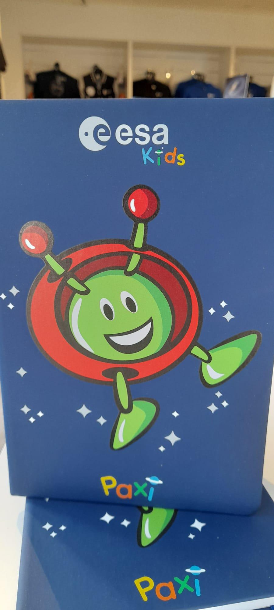 A photo showing an ESA Paxi Notebook. The notebook is blue and has a drawing of a green alien with antennae on the front. Paxi is a mascot for ESA kids.